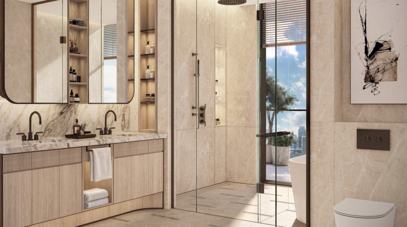 The luxurious bathroom in the Atelis Design District features beige marble walls and floor, a large glass-enclosed shower and double washbasin with marble countertop. Sunlight filters through a window, giving glimpses of blue sky and trees. Elegant decor, including a modern abstract painting, enhances the space.