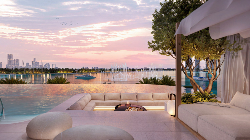 Luxurious outdoor relaxation area with comfortable sunken seating overlooking an infinity pool. Beyond, a peaceful lake with the Atelis Design District skyline in the distance at sunset. Decorative trees and subdued lighting accentuate the serene ambience.