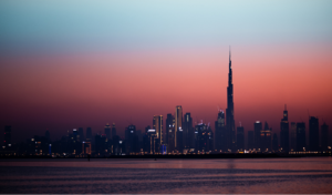 Silhouette d'une ville au crépuscule, avec sa haute tour imposante rappelant Dubaï. Le ciel passe d'un bleu profond à un dégradé chaud d'orange et de rose. Au premier plan, l'eau calme reflète les lumières vibrantes de la ville et laisse deviner ses 4 millions d'habitants.