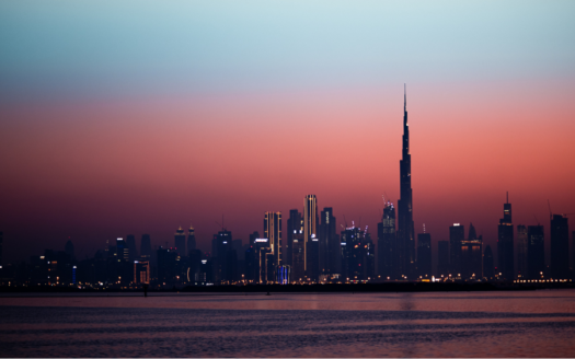 Silhouette einer Stadt in der Abenddämmerung mit einem hohen, imposanten Turm, der an Dubai erinnert. Der Himmel wechselt von einem tiefen Blau in eine warme Abstufung von Orange und Rosa. Im Vordergrund spiegelt das ruhige Wasser die pulsierenden Lichter der Stadt wider und lässt ihre 4 Millionen Einwohner erahnen.