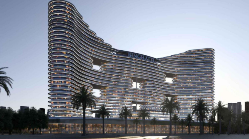 Un moderno rascacielos curvo, el "Binghatti Aquarise", presenta ventanas iluminadas y un diseño ondulante, enmarcado por palmeras al atardecer.