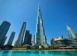 Das Bild hebt den Burj Khalifa, einen imposanten Wolkenkratzer in Dubai, vor einem azurblauen Himmel hervor. Die umliegenden Gebäude und ein friedliches Gewässer sublimieren die Landschaft und rufen ein Gefühl von Sicherheit und Gelassenheit hervor.