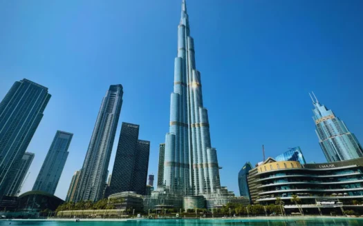 The image highlights the Burj Khalifa, an imposing Dubai skyscraper, standing out against an azure blue sky. The surrounding buildings and a tranquil body of water enhance the landscape, evoking a sense of security and serenity.