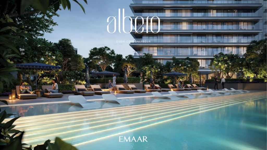 Una lussuosa area piscina all'aperto al tramonto, con moderni lettini parzialmente sommersi. Alti edifici e vegetazione lussureggiante circondano la piscina. Le scritte "albeiro" e "Emaar" sono visibili in alto e in basso nell'immagine.