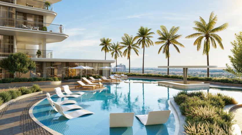 Modern outdoor pool area in a luxury building with sun loungers in and around the water, palm trees, greenery and a cityscape visible in the distance under a clear sky.