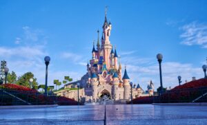 Une vue grand angle du château rose et bleu de la Belle au bois dormant à Disneyland Abu Dhabi, avec un ciel bleu clair et des jardins paysagers avec des fleurs rouges de chaque côté du chemin d'entrée.