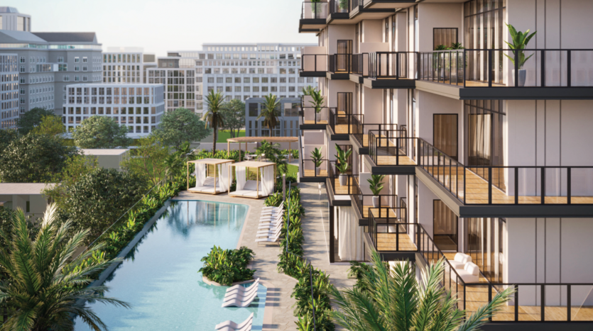 Un moderno edificio di appartamenti con balconi si affaccia su una piscina fiancheggiata da lettini, cabanas, palme e verde; sullo sfondo si vedono il DoubleTree by Hilton Dubai e gli edifici della città.