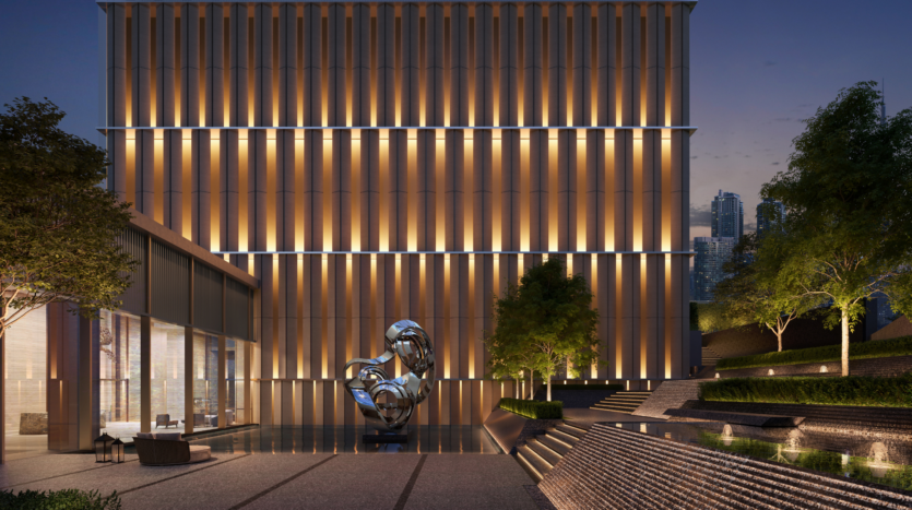 Edificio moderno iluminado verticalmente al atardecer, con una escultura abstracta reflectante, espacio exterior, árboles y paisaje de varios niveles con elementos acuáticos en primer plano. Al fondo se ven los rascacielos de la ciudad.