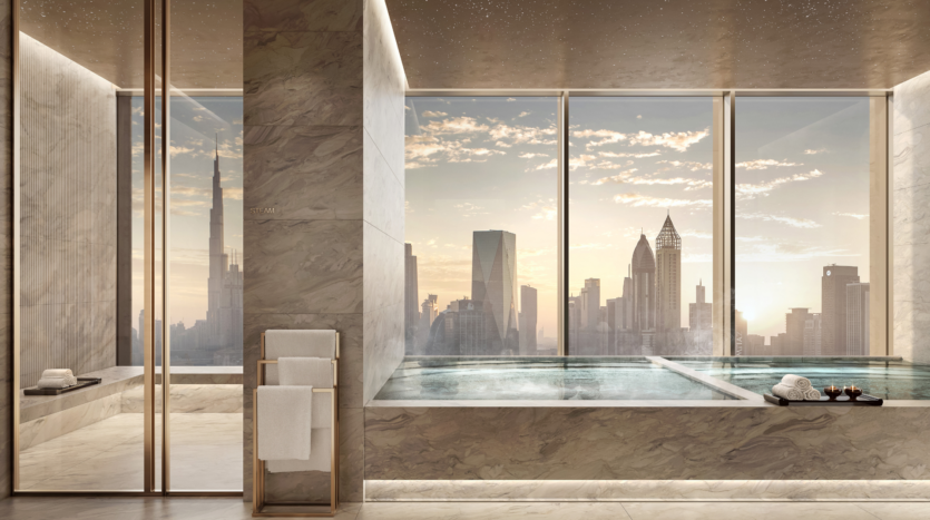 Lujoso cuarto de baño de mármol con una gran bañera junto a ventanales que ofrecen impresionantes vistas de la ciudad al atardecer. Junto a la bañera hay un toallero y toallas enrolladas.
