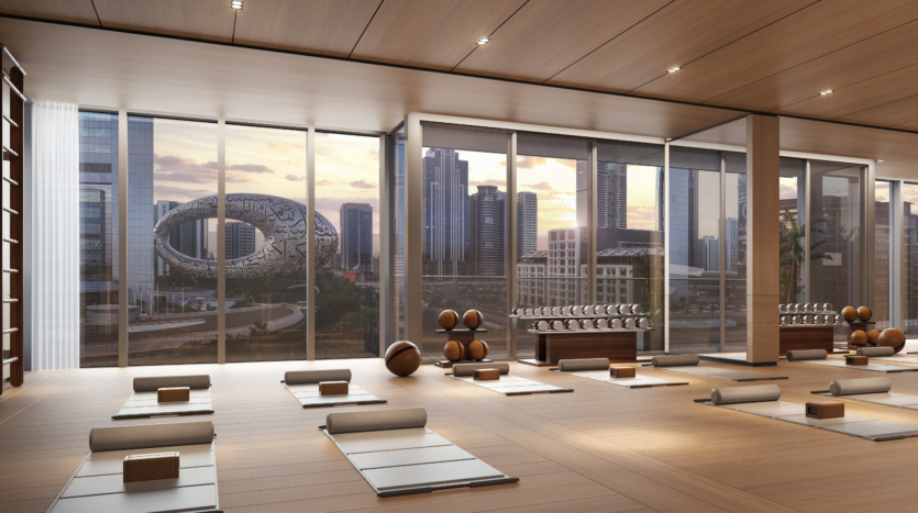 Un moderno estudio de yoga con esterillas, almohadones y bloques de yoga cuidadosamente dispuestos, con ventanales con vistas a los tejados de la ciudad y al Museo del Futuro de Dubai al atardecer.