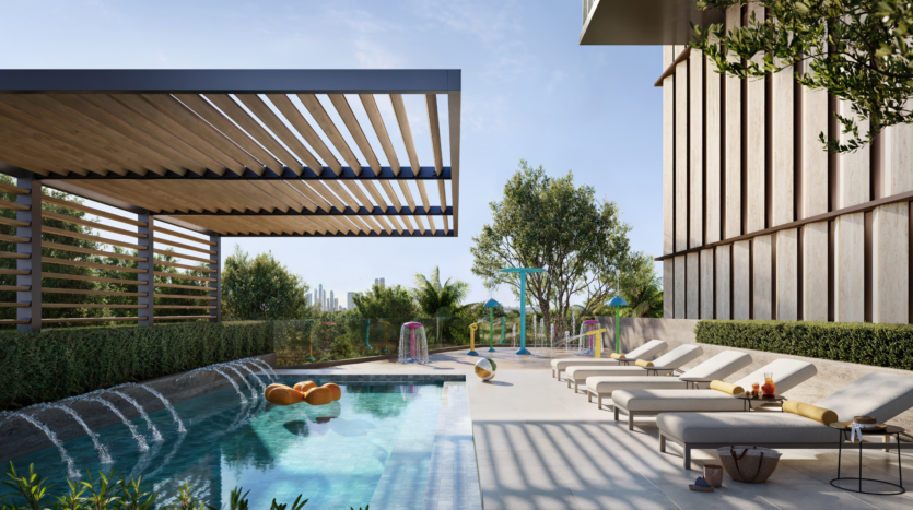 Moderna zona de piscina al aire libre con juegos de agua, tumbonas, pérgola con sombra, árboles y jardines. Coloridas estructuras de juego y un horizonte urbano visible al fondo bajo un cielo azul celeste.