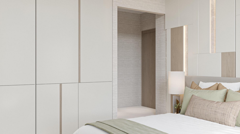 Minimalist room at Provenza Residences Dubai with light-colored walls, built-in wardrobes, a carefully made bed with pastel green and beige pillows and a bedside lamp next to an open door leading to a corridor.