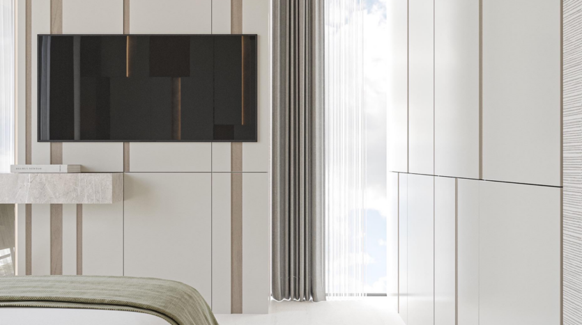 Minimalist room at Provenza Residences Dubai, featuring a wall-mounted flat-screen TV, light-colored paneled walls, a small floating bookshelf, floor-to-ceiling curtains and a bed with a green blanket in the foreground.