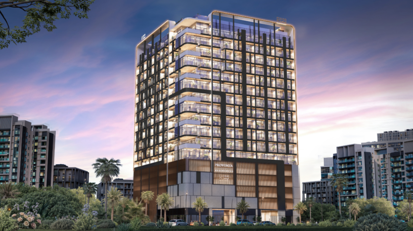 A modern, multi-storey glass building with illuminated balconies stands among palm trees and greenery at sunset, surrounded by other contemporary buildings - an iconic view from Provenza Residences Dubai.