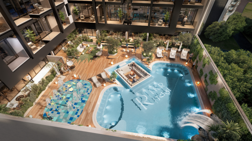 Aerial view of a luxury outdoor pool at Provenza Residences Dubai, featuring a fountain, poolside loungers, green plants, cabanas and "RIVA" written at the bottom of the pool, surrounded by modern apartment buildings and trees.