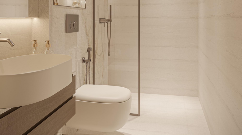 The modern, minimalist bathroom at Provenza Residences Dubai features beige tiles, a wall-mounted toilet, a round sink on a wooden vanity, two soap dispensers and a glass-enclosed shower with rainfall showerhead.
