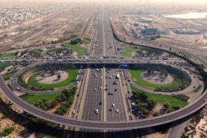 Vue aérienne d'un grand échangeur routier à plusieurs voies et viaducs, avec l'aménagement d'Umm Suqeim, des espaces verts paysagers, un trafic intense et un paysage urbain tentaculaire en arrière-plan.