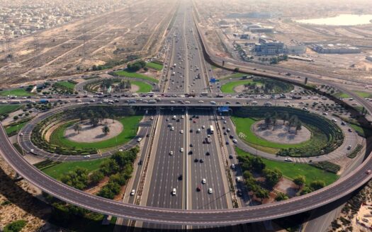 Luftaufnahme eines großen Verkehrsknotenpunkts mit mehreren Fahrspuren und Viadukten, mit der Umm Suqeim-Entwicklung, landschaftlich gestalteten Grünflächen, starkem Verkehr und einer tentakelartigen Stadtlandschaft im Hintergrund.