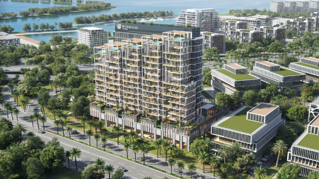 Vista aerea di un moderno edificio multipiano con terrazze sul tetto e verde, circondato da palme, strade e altre strutture contemporanee vicino all'acqua.