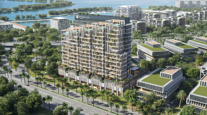 Vista aerea di un moderno edificio multipiano con terrazze sul tetto e verde, circondato da palme, strade e altre strutture contemporanee vicino all'acqua.