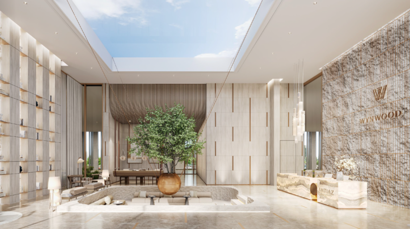 Una moderna hall d'albergo con soffitti alti, un grande lucernario, un'area lounge centrale incassata attorno a un albero in vaso, pavimenti in marmo e un banco della reception con un'insegna "WYNWOOD" sulla parete in pietra.