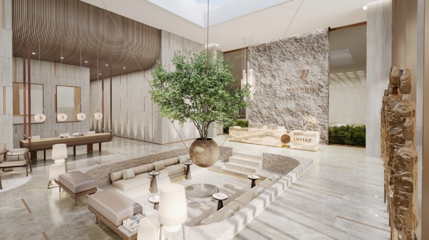 Una hall spaziosa e moderna, dai toni neutri, con un grande albero in vaso come elemento centrale, un'area lounge incassata, un bancone della reception e tocchi di pietra naturale. I lucernari lasciano entrare la luce naturale, creando un'atmosfera ariosa.