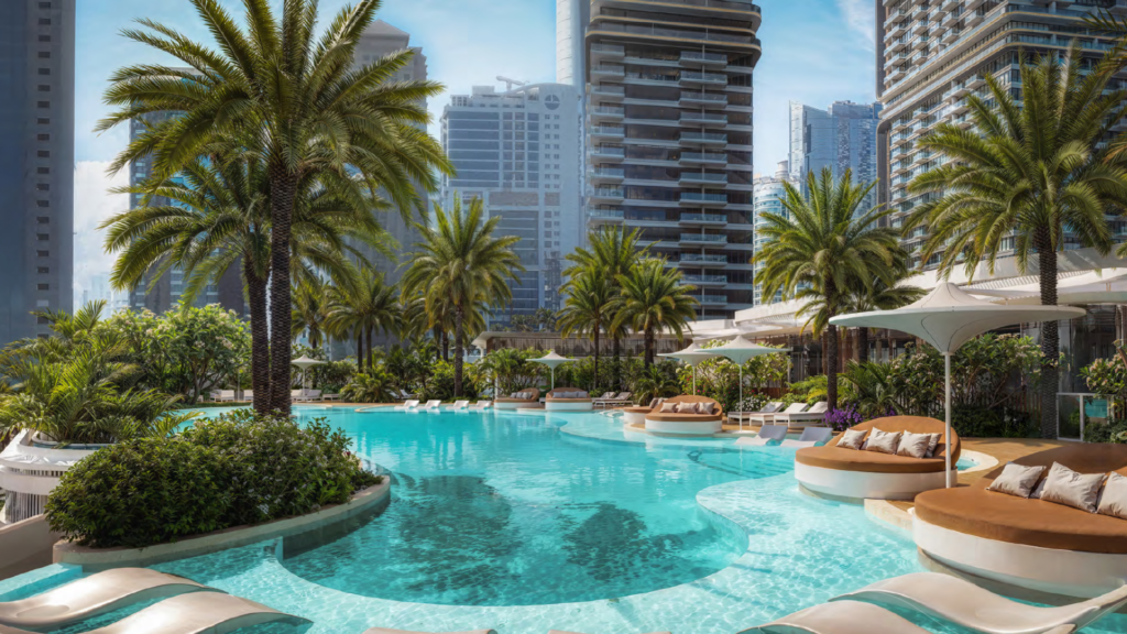 Luxury outdoor pool surrounded by palm trees, sun loungers and modern high-rise buildings in an urban setting under a clear blue sky.