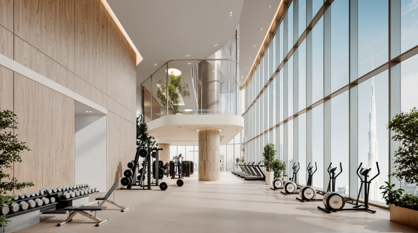 Bright, modern gym with large picture windows, potted plants, weight racks, benches, treadmills and exercise bikes lined up along the glass wall; a view of the city's rooftops is visible outside.