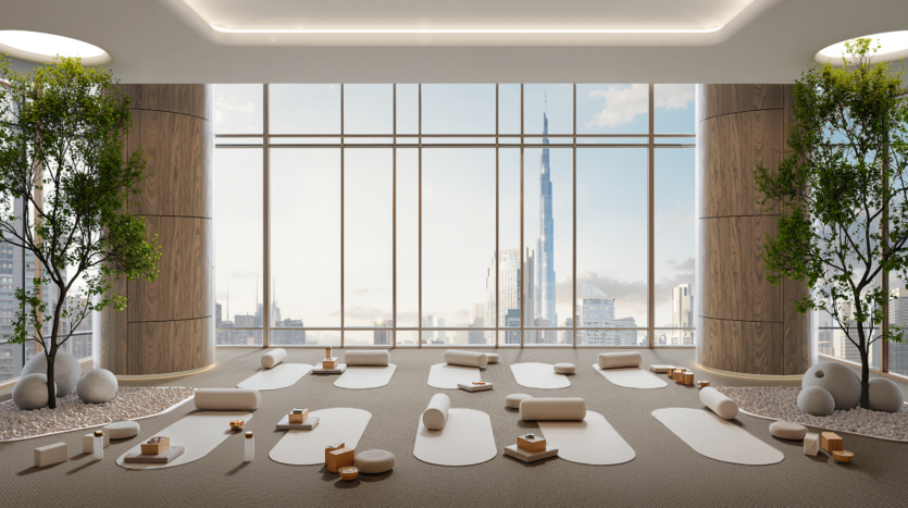A spacious, modern yoga studio with large picture windows overlooking the city, including a skyscraper. The room is equipped with yoga mats, bolsters and potted trees, creating a serene atmosphere.