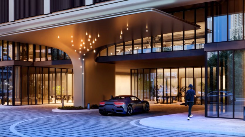 The entrance to a modern building features large bay windows, warm lighting and unique ceiling fixtures. A luxury sports car is parked in front of the entrance while a person makes his way to the doors.