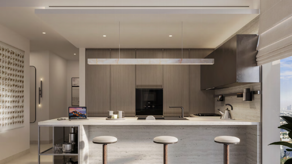 Modern kitchen in neutral tones featuring a large island with three white bar stools, built-in cooktop, stylish oven, dark cabinets, kettle, laptop and large window with city views.