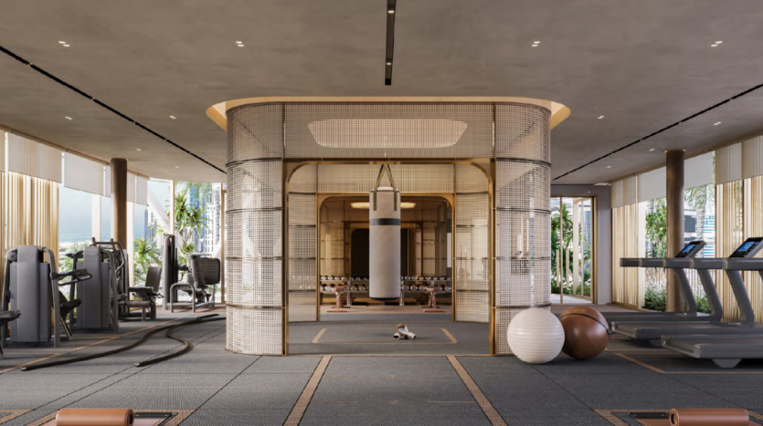 Modern, spacious gym with large windows, featuring a central boxing area equipped with a hanging punching bag, surrounded by weight machines, free weights and fitness balls. The room is bathed in natural light and the greenery is visible outside.