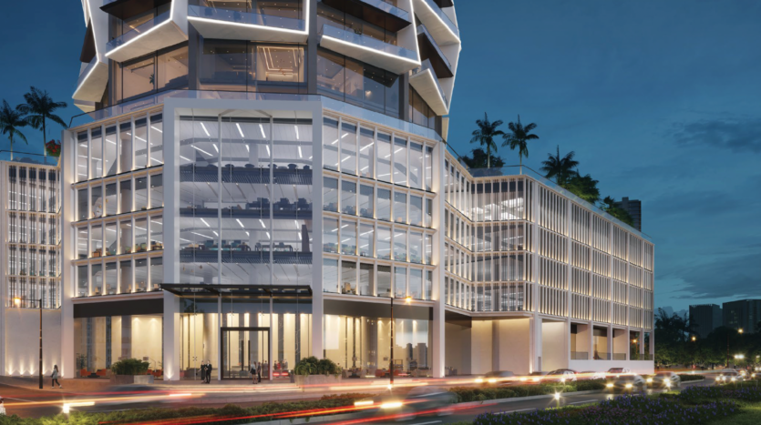 A modern glass office building is lit up at dusk, with geometric architecture, large windows revealing illuminated interiors, palm trees on the terraces and blurred car headlights on the road ahead.