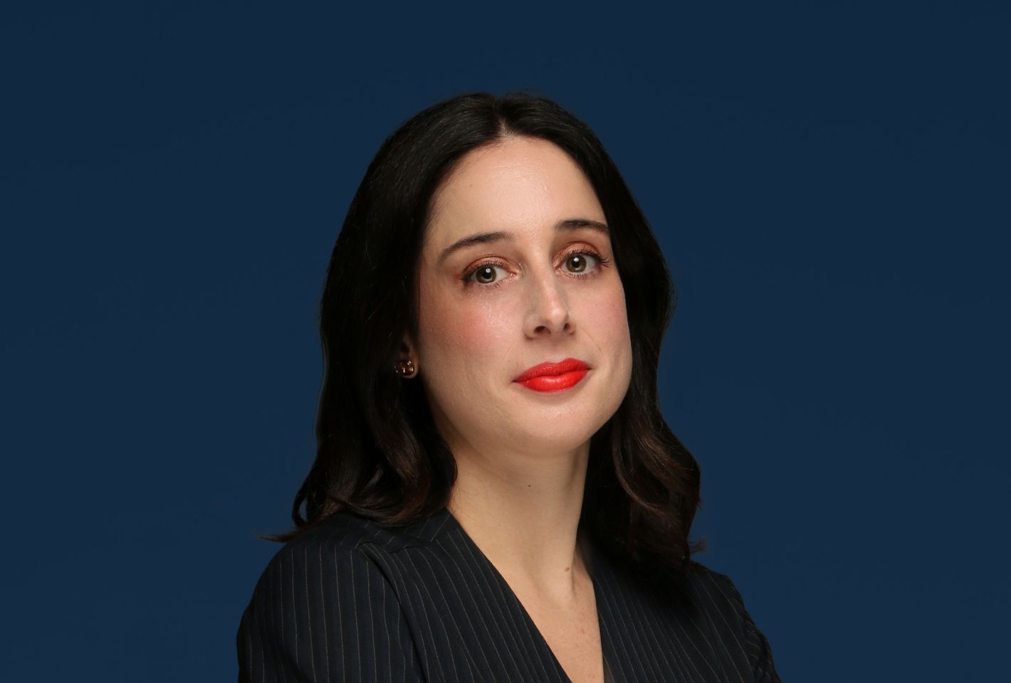 Frau mit halblangem braunem Haar in einem dunkel gestreiften Blazer, die mit neutralem Gesichtsausdruck und rotem Lippenstift in die Kamera blickt, vor einem blauen Hintergrund - spiegelt das professionelle Selbstbewusstsein auf dem Immobilienmarkt in Dubai wider.