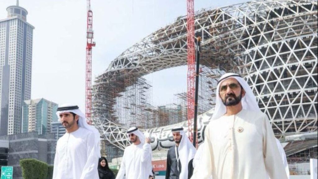 Un groupe d'hommes portant des vêtements traditionnels émiratis sortent devant un grand bâtiment circulaire futuriste en construction avec des grues, entouré de gratte-ciel.