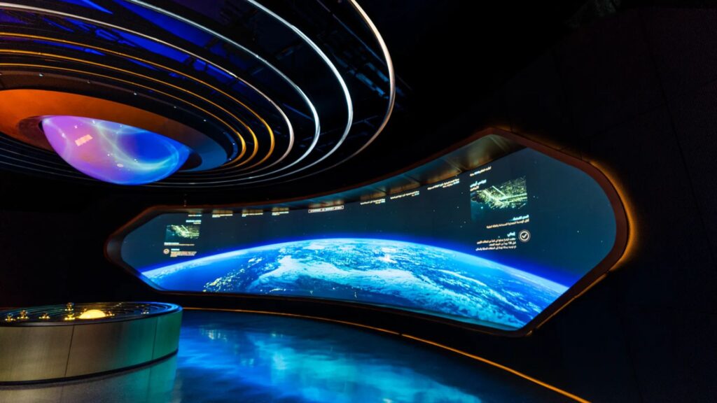 Salle futuriste avec un grand écran incurvé affichant une vue de la Terre depuis l'espace, éclairé par des lumières bleues et jaunes, et une console circulaire au premier plan.