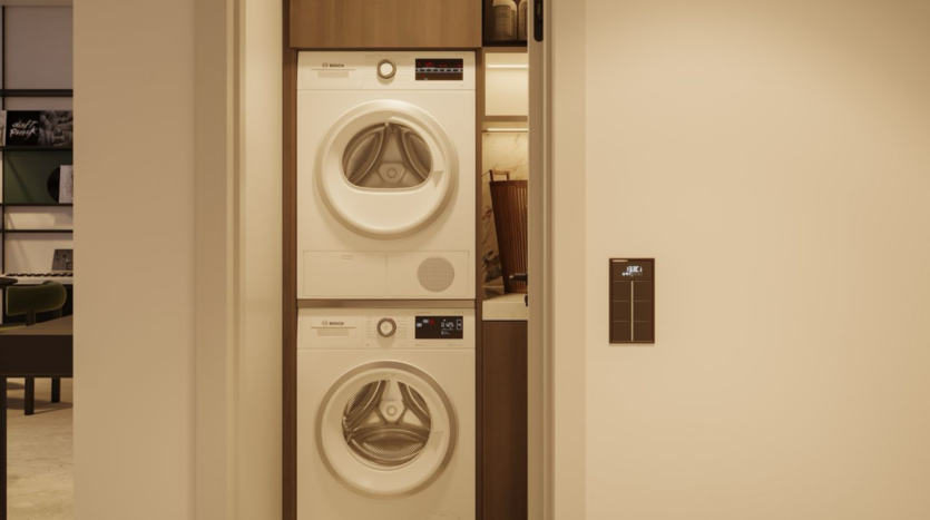 A stacked washer and dryer set is installed in a small linen closet with a sliding door, adjacent to a modern living space. The appliances are white and shelves are visible above them.