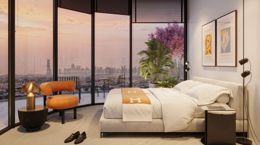 Modern bedroom with floor-to-ceiling windows overlooking a city at sunset, plush bed with beige blanket, orange chair, bedside tables, framed art on the wall, potted plant and shoes on the floor.
