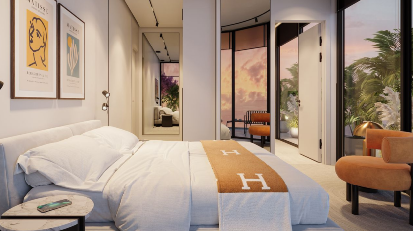 Modern bedroom with well-made bed, beige blanket and white pillows. Large windows reveal a brilliant sunset and greenery outside. Minimalist furnishings include chairs, a side table with a telephone and artwork on the walls.