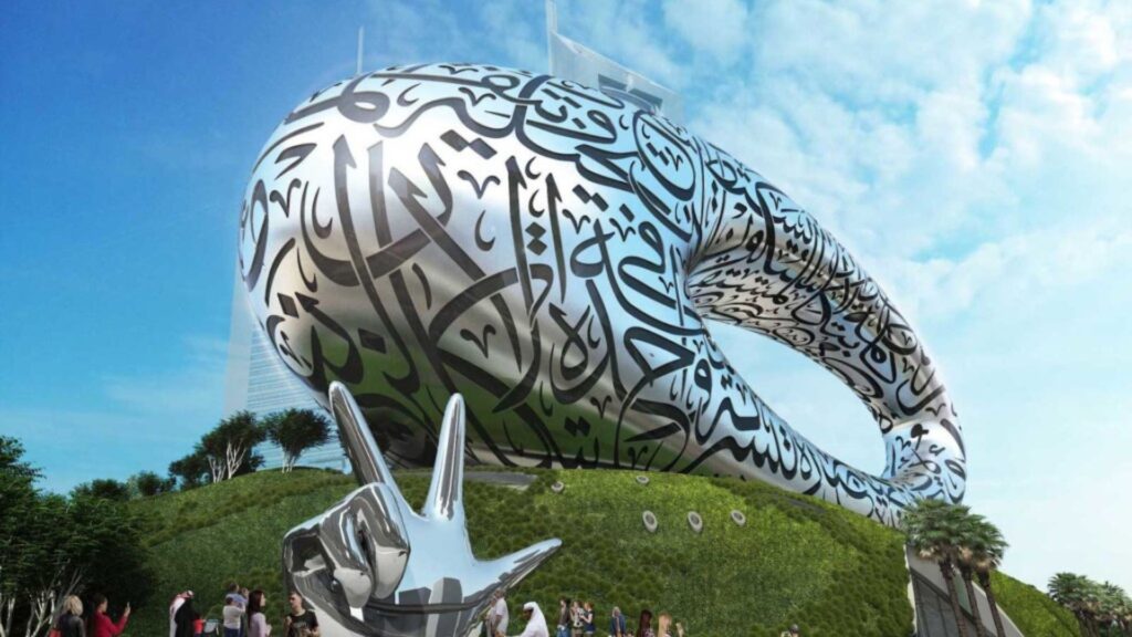 Un bâtiment futuriste de forme ovale, couvert de calligraphie arabe, se dresse au sommet d'une colline verdoyante, sous un ciel bleu. Des personnes se rassemblent près d'une sculpture argentée brillante représentant une main faisant un signe de paix au premier plan.