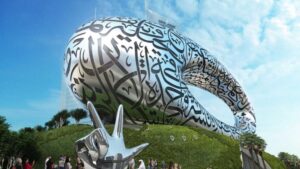 Ein futuristisches, ovales Gebäude, das mit arabischer Kalligraphie bedeckt ist, erhebt sich unter blauem Himmel auf einem grünen Hügel. Menschen versammeln sich neben einer glänzenden silbernen Skulptur, die eine Hand darstellt, die im Vordergrund ein Zeichen des Friedens macht.
