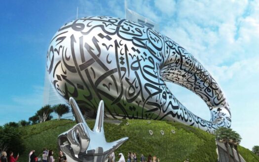 Ein futuristisches, ovales Gebäude, das mit arabischer Kalligraphie bedeckt ist, erhebt sich unter blauem Himmel auf einem grünen Hügel. Menschen versammeln sich neben einer glänzenden silbernen Skulptur, die eine Hand darstellt, die im Vordergrund ein Zeichen des Friedens macht.