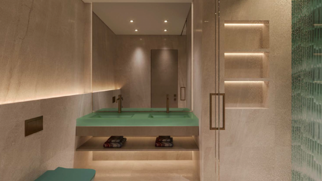 Modern bathroom with beige stone walls and floor, large green double sink, two faucets, glass shower door, built-in shelves, recessed lighting and neatly folded towels stored under the sink.