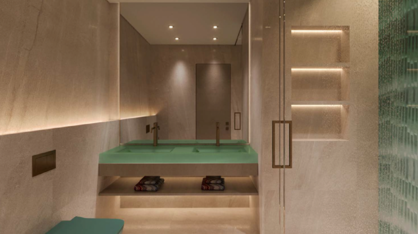 Modern bathroom with beige stone walls, a large green double washbasin and subdued recessed lighting, inspired by the elegance of Dubai's Octa Isle. Built-in shelves store folded towels, and a glass shower is visible on the right.