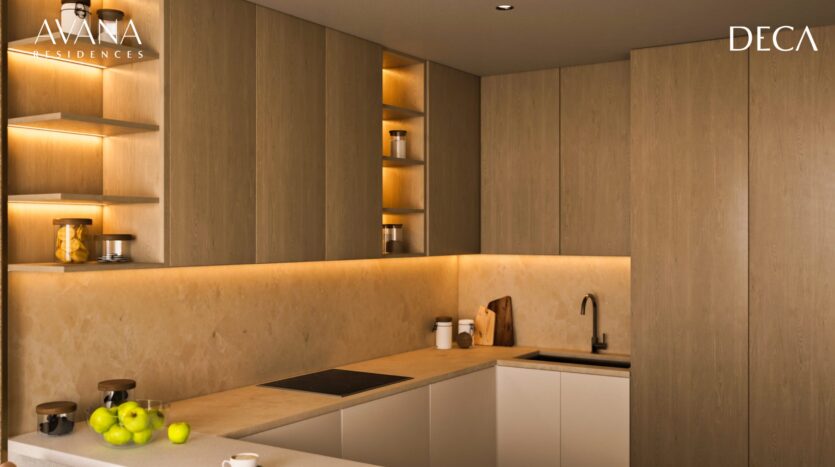 Modern kitchen in Avana residences by Deca Properties in Dubai, featuring light wood cabinets, open shelves with jars and fruit, beige marble backsplash and countertop, built-in stove, under-cabinet lighting; "AVANA" and "DECA" logos in upper corners.