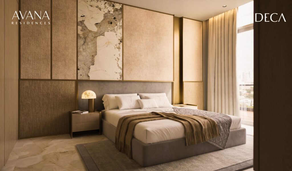 A modern, sunny bedroom with neutral tones, a king-size bed with gray and beige bedding, a bedside table with lamp, floor-to-ceiling windows and paneled wall decoration. The logos "AVANA Residences by Deca Properties in Dubai" and "DECA" appear.
