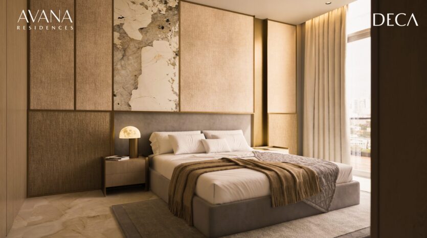 A modern, sunny bedroom with neutral tones, a king-size bed with gray and beige bedding, a bedside table with lamp, floor-to-ceiling windows and paneled wall decoration. The logos "AVANA Residences by Deca Properties in Dubai" and "DECA" appear.