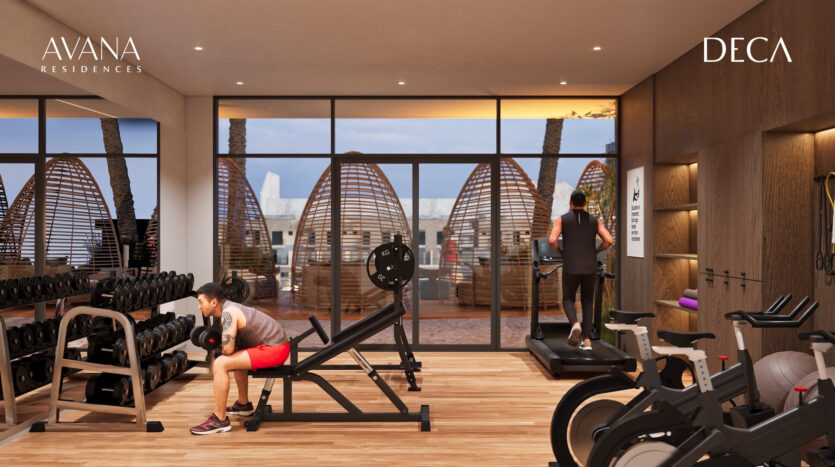 The modern gym at Avana Residences by Deca Properties in Dubai features large windows, weight-lifting equipment, treadmills and exercise bikes. Two people work out while cabanas and palm trees can be seen outside.