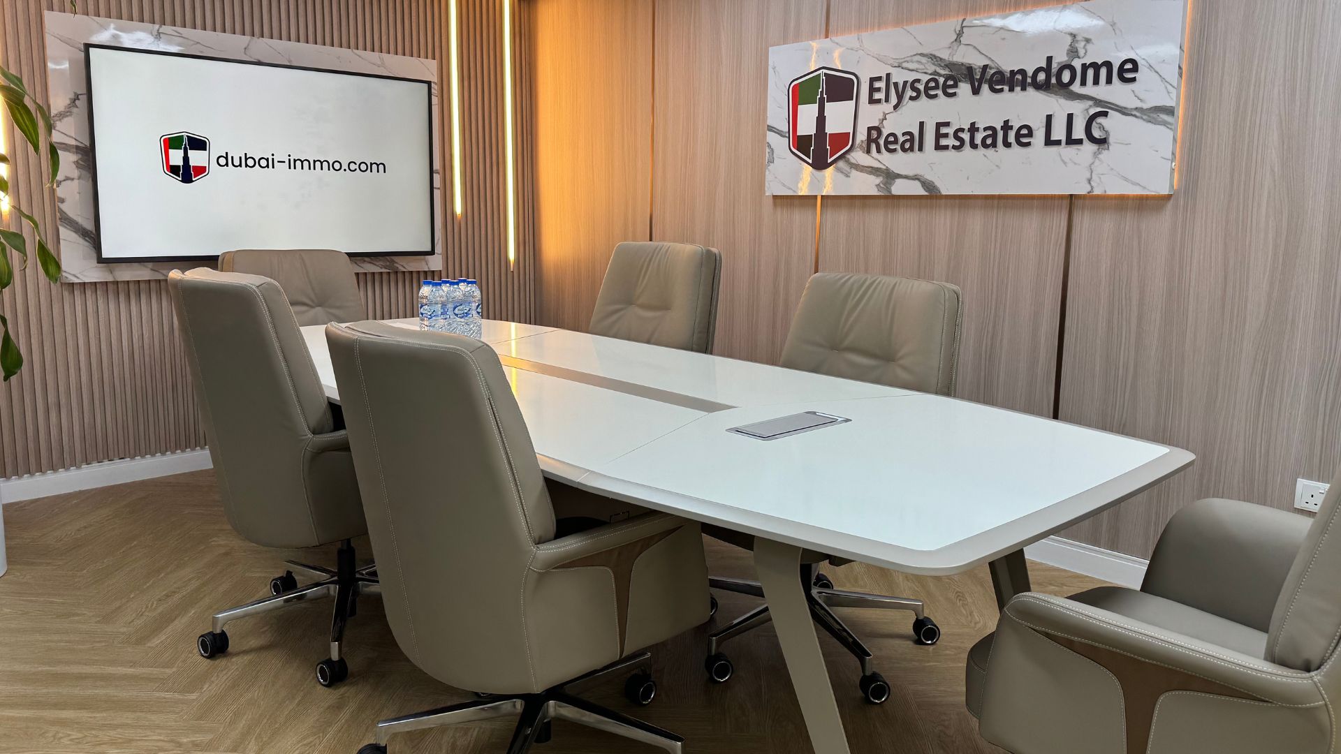 Una moderna sala conferenze con un tavolo bianco, sei sedie beige, una bottiglia d'acqua, una pianta e le scritte "Elysee Vendome Real Estate LLC" e "Dubai Immo" alle pareti. Lo spazio ha pavimenti in legno e un'illuminazione calda.