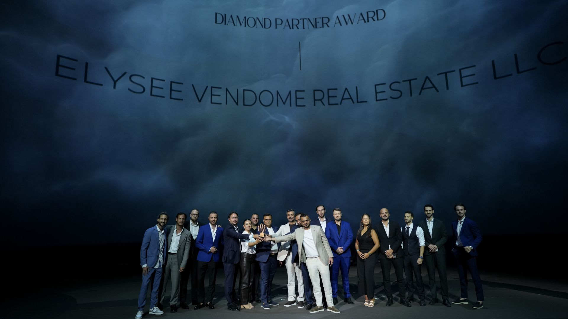 Un folto gruppo in abiti formali si trova sul palco sotto un cartello che recita "Diamond Partner Award, Elysee Vendome Real Estate LLC". Posano con un trofeo che rappresenta l'eccellenza di Dubai Immo su un drammatico sfondo nero.